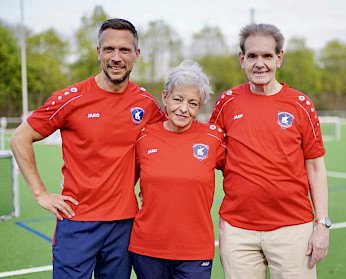 Der erste und zweite Vorsitzende halten die Kassenwartin im Arm. Im Hintergrund ist der Fußballplatz vom Störtebeker SV verschwommen zu erkennen. Der erste und zweite Vorsitzende halten die Kassenwartin im Arm. Im Hintergrund ist der Fußballplatz vom Störtebeker SV verschwommen zu erkennen.
